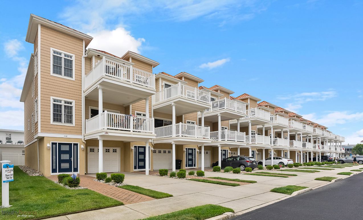 Row of beige townhouses with white balconies and garages, under blue skies. They evoke a calm, suburban feel with well-kept lawns and driveways.